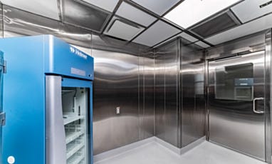 A cleanroom storage or preparation area with a Helmer medical-grade refrigerator and stainless steel door with observation window