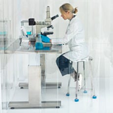 Female laboratory scientist in white coat and blue gloves sitting on tall stainless steel lab stool while using microscope