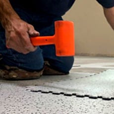 Installation of StaticWorx GroundSafe ESD's interlocking floor tiles using an orange rubber mallet, pressing tiles into place on a speckled gray floor surface